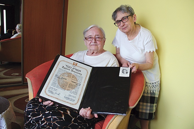 ▲	Krystyna pokazuje dyplom Sprawiedliwi wśród Narodów Świata przyznany jej rodzicom. Za nią jej córka Danuta.