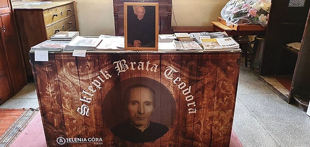 Stół z prasą katolicką i książkami wieńczy dibond z wizerunkiem zmarłego pijara.