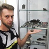 ◄	Sebastian Mielczarek obok półki z przedmiotami znalezionymi na terenie dawnego obozu jenieckiego. Na dłoni trzyma fragment zapalnika artyleryjskiego.