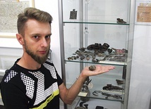 ◄	Sebastian Mielczarek obok półki z przedmiotami znalezionymi na terenie dawnego obozu jenieckiego. Na dłoni trzyma fragment zapalnika artyleryjskiego.