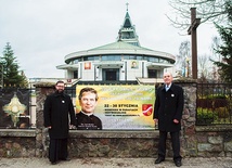 ▲	Ks. Rafał Burnicki SDB i Jan Klawiter z banerem zachęcającym do udziału w nowennie do świętego przed rumskim sanktuarium.